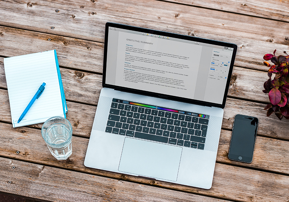 A document is open on a laptop, with a phone, a notebook and pen, a glass of water and a plant around the laptop, all sitting on a table 